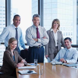 A diverse group of five business professionals in formal attire are smiling around a conference table
