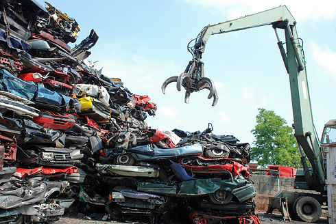 A large claw crane lifts scrap metal near a huge mound of stacked, wrecked cars