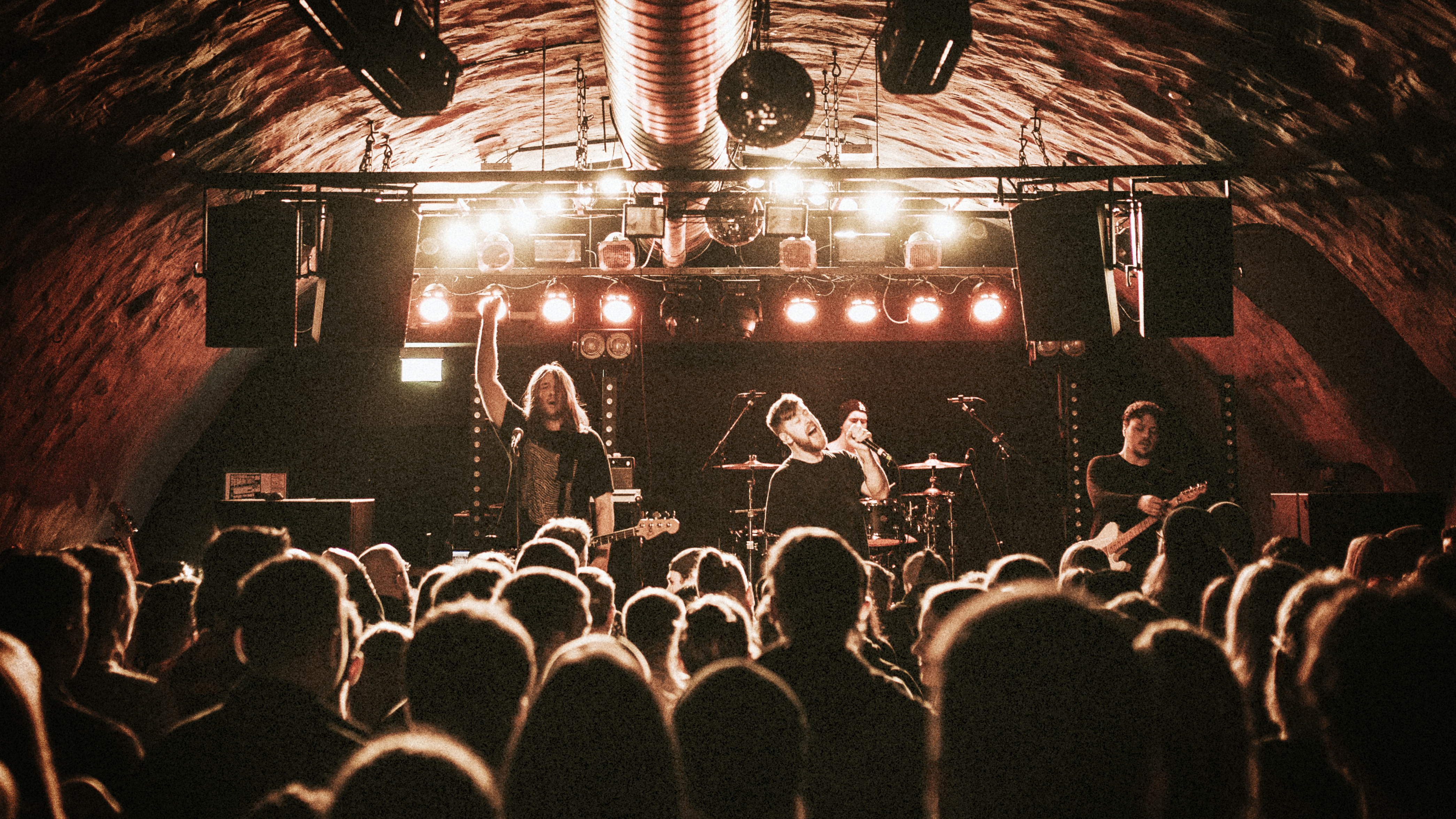 Band performing live on stage in a warm-lit venue with an active audience.