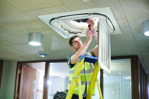 A technician in a high-visibility vest stands on a ladder, repairing an overhead air conditioning unit