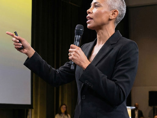 Person in a black suit speaks into a microphone, gesturing with a hand. Yellow-lit stage with an audience in the background.