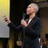Person in a black suit speaks into a microphone, gesturing with a hand. Yellow-lit stage with an audience in the background.