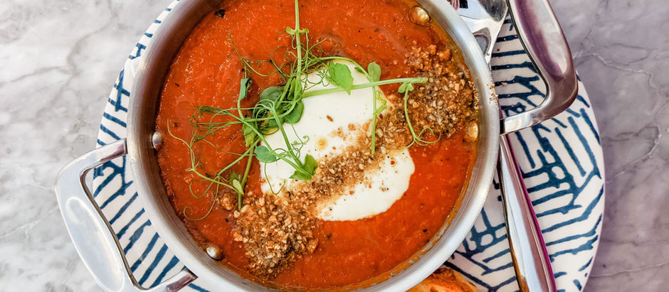 A bowl of smooth tomato soup topped with a swirl of sour cream in the center, scattered rye crumbs across the top, and delicate green herbs for garnish.