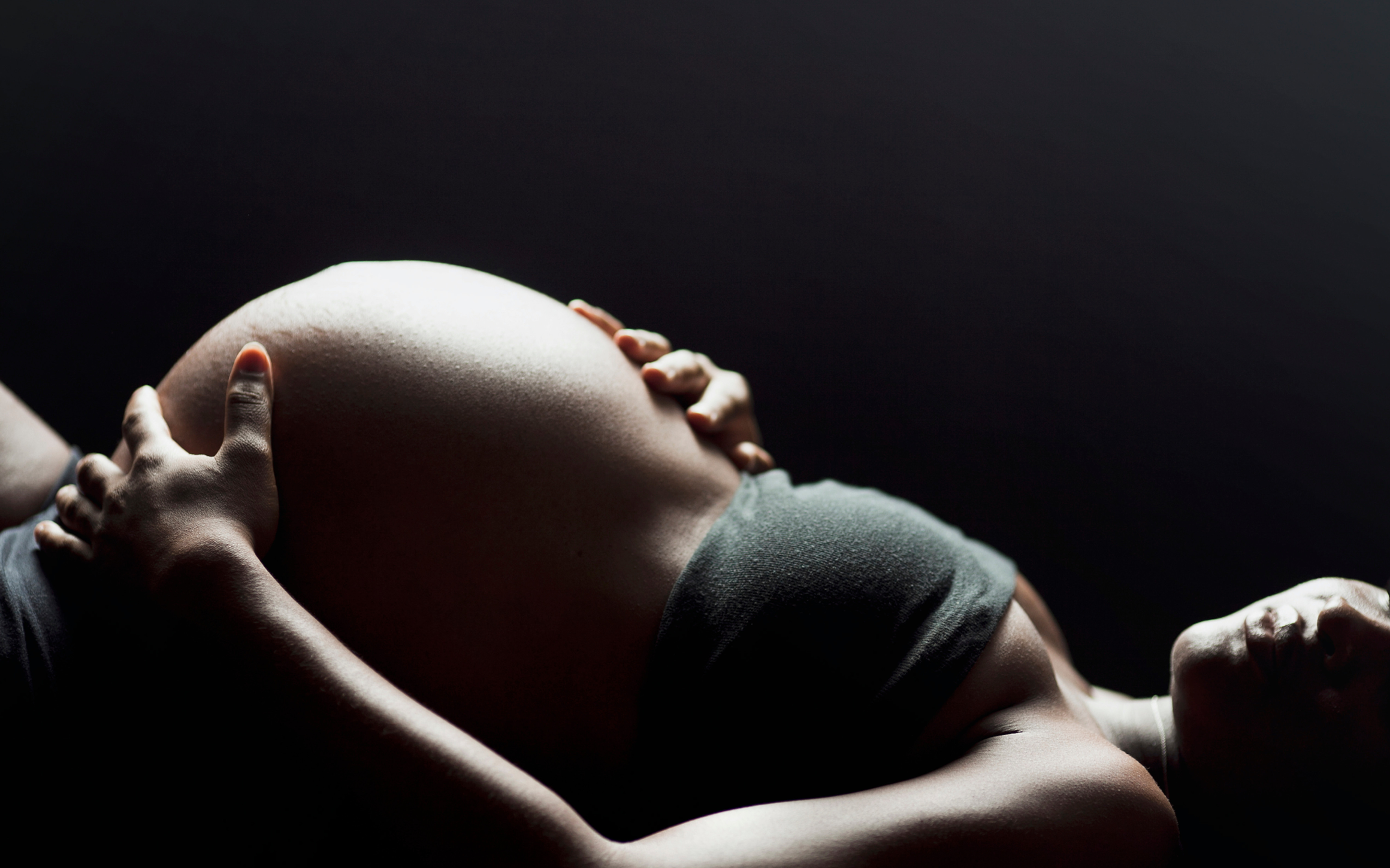 A woman rests, her hands gently cradling her pregnant belly, illuminated against a dark background.