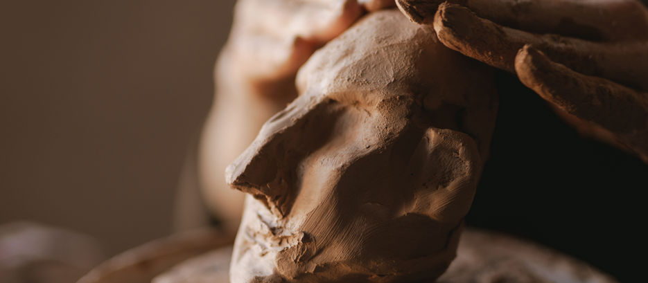 Mãos modelam escultura de barro em forma de rosto. Fundo desfocado. Tons terrosos e textura detalhada evidenciam processo criativo.
