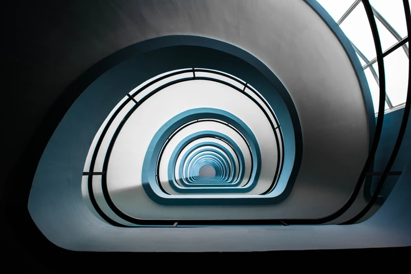 A spiral staircase seen from below looking upward through many floors, bathed in natural light with soft blue tones, representing the ongoing and layered journey of personal growth and adaptation in the Identity, Change and Adaptation Series at Availia.