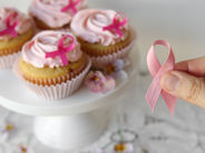 Hand holding a pink awareness ribbon beside cupcakes topped with pink ribbon icing on a white stand
