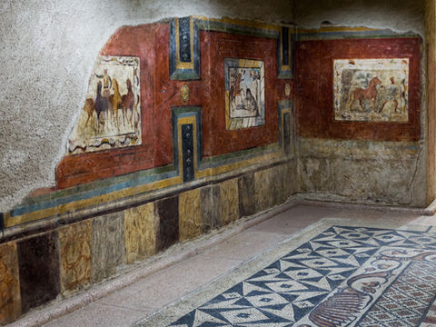 Mur antique orné de fresques romaines colorées et d’un sol en mosaïque, dans un couloir de pierre aux tons rouge et ocre.