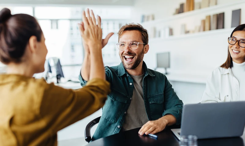 Two coworkers high-five during a work meeting while a third colleague smiles.
