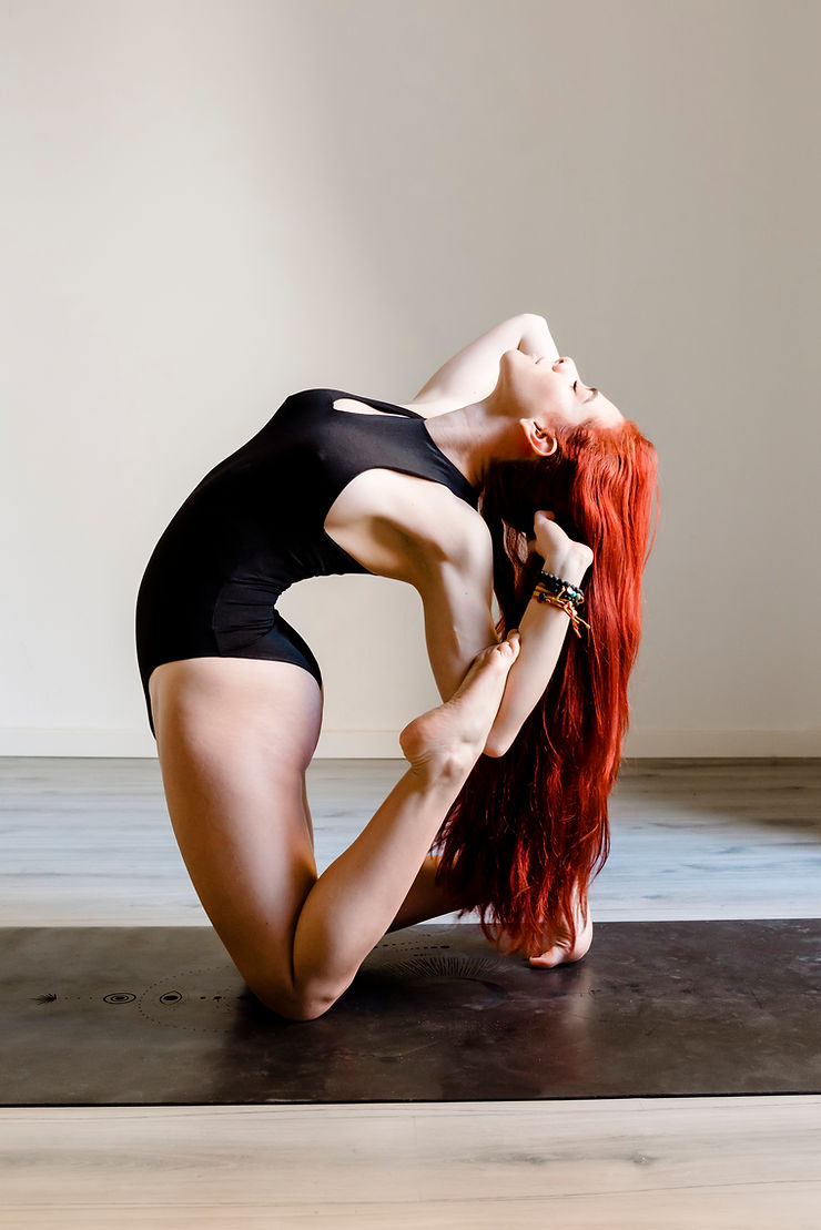 Woman in black leotard doing yoga on a mat, arching back with long red hair flowing. Calm mood in a minimalist room.