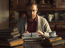 A man sitting at a typewriter. He's wearing glasses and a cozy sweater and looks to be in an older home, surrounded by books.
