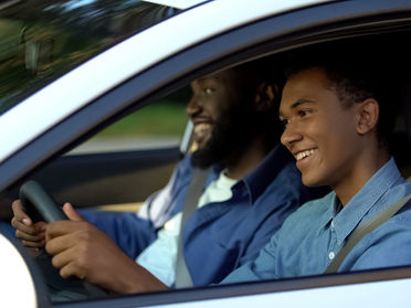 Two people in a car, smiling. The driver is focused on the road, wearing a blue shirt. The setting is outdoors, captured from the side. Connecticut auto insurance