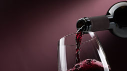 Red wine being poured from a bottle into a clear wine glass against a rich burgundy background, with frothy bubbles forming on the surface.