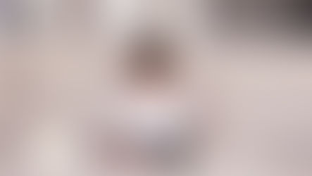 Toddler reading a book on a beige sofa, surrounded by stacked books and a teddy bear. The room is cozy with a soft, neutral tone.