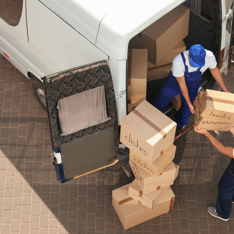 Two movers in blue overalls and white shirts are loading labeled cardboard moving boxes into the back of a white moving van. One box being handed up is labeled "books Moving." Other boxes are labeled "dishes" and "kitchen." The photo is taken from a high angle, looking down onto a paved brick surface.