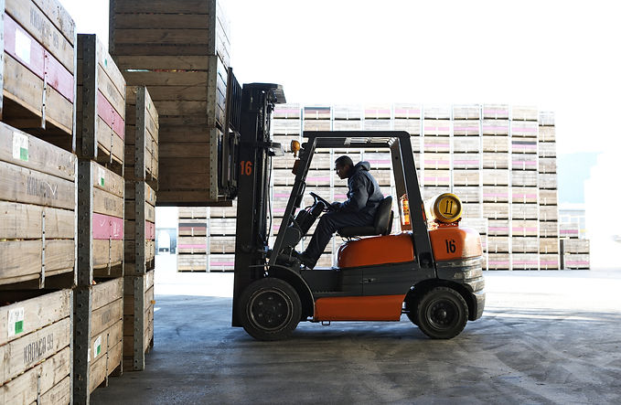 A worker in a dark hoodie operating an orange forklift, stacking wooden crates in an outdoor storage yard