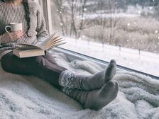 Cozy scene of a person in a sweater and patterned socks, reading a book by a window. Snow falls outside, creating a peaceful winter mood.