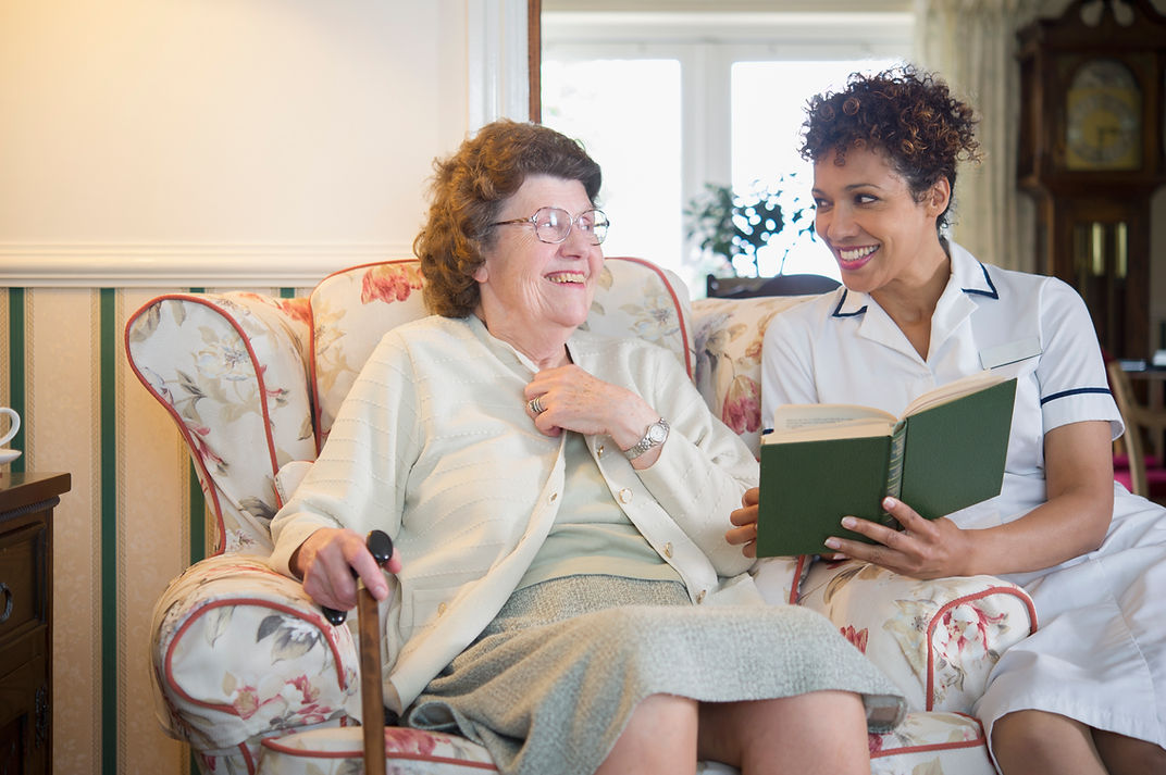 A smiling elderly woman and a caregiver share a joyful moment on a floral sofa