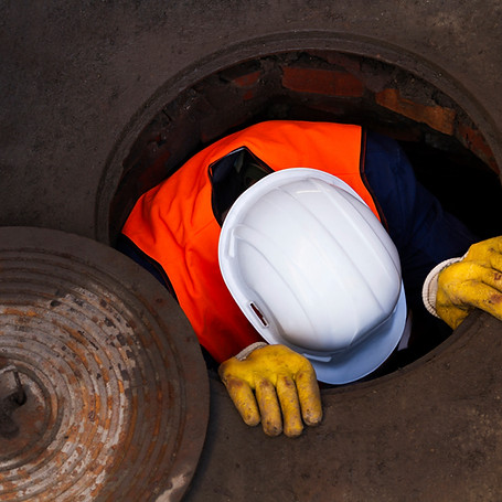 Worker wearing a white hard hat and orange vest climbs into a manhole