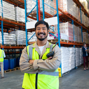 Smiling worker in a yellow vest holds a scanner in a warehouse. Background shows shelves of goods and colleagues having a discussion.