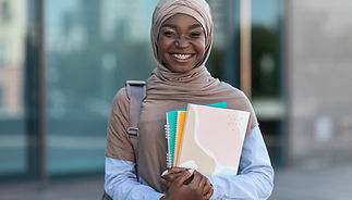 A smiling young woman wearing a beige hijab and braces holds several notebooks and a backpack strap over her shoulder. She stands outdoors in front of a modern building, exuding confidence and readiness for her physiotherapy training. Her bright smile reflects the excitement and dedication she brings to advancing her skills through professional development courses.