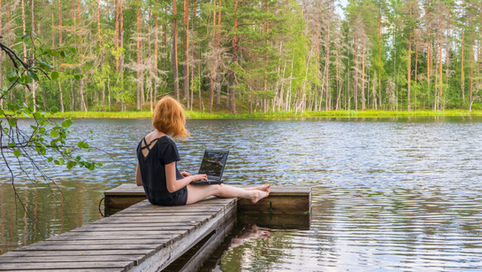 A woman with red hair sits on a wooden dock by a tranquil lake, working on a laptop. She is surrounded by lush greenery and tall trees, reflecting the serene, flexible lifestyle often associated with freelancers.