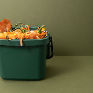 Green compost bin with vegetable peels and food scraps against an olive background, evoking an eco-friendly, waste-recycling theme.