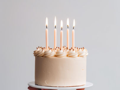 Hand holds a white cake with five lit candles against a plain gray background. The cake has creamy swirls and creates a celebratory mood.