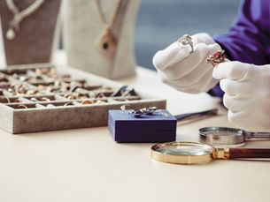 White-gloved hands examine two rings with a magnifying glass nearby. A jewelry box and necklace stand in the background. Elegant setting.