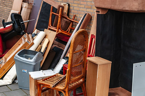 Piles of old wooden chairs and household waste discarded outside a house next to a grey wheelie bin