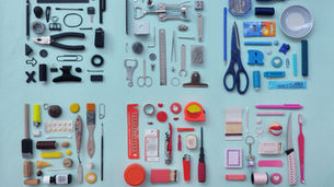 Assorted objects arranged by color on a light blue background, featuring tools, office supplies, and household items in black, silver, blue, yellow, red, and pink.