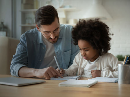 A father helping his young child with writing homework at a table at home.