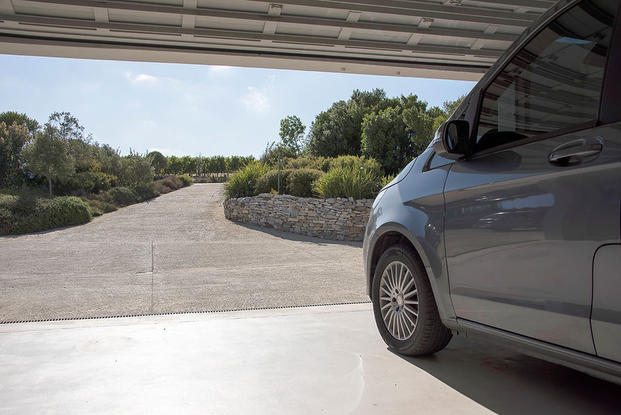 A gray car is parked in a garage with an open door, overlooking a gravel driveway