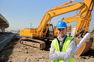 Construction worker in blue hard hat and yellow vest standing with arms crossed in front of large excavators at site
