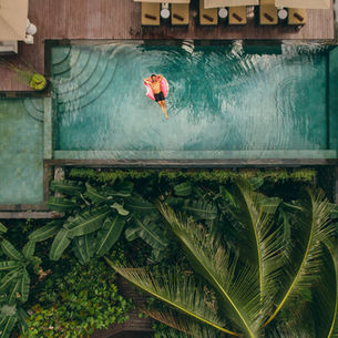 Person in a pink float relaxes in a turquoise pool surrounded by lush greenery and wooden deck chairs, creating a serene, tropical vibe.