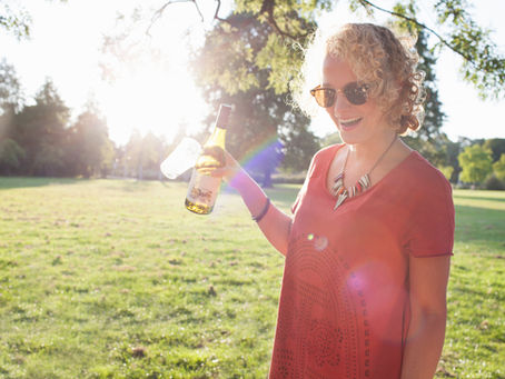 Vrouw in rood shirt lacht en houdt een fles wijn vast in een zonnig park. Ze draagt een zonnebril en ketting. Lenteachtige sfeer.