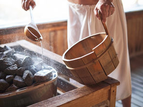 Person pouring water from a ladle onto hot sauna stones, creating steam. Wooden bucket in hand, warm wooden interior setting.