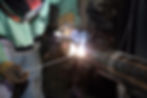 A fabricator welding a large steel pipe using a welding rod, with bright sparks and smoke