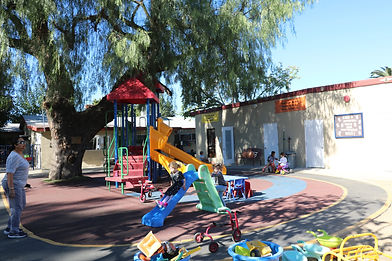 A sunny preschool playground with a colorful slide structure and scattered ride-on toys, bordered by a school building where a few adults and children sit in the shade.