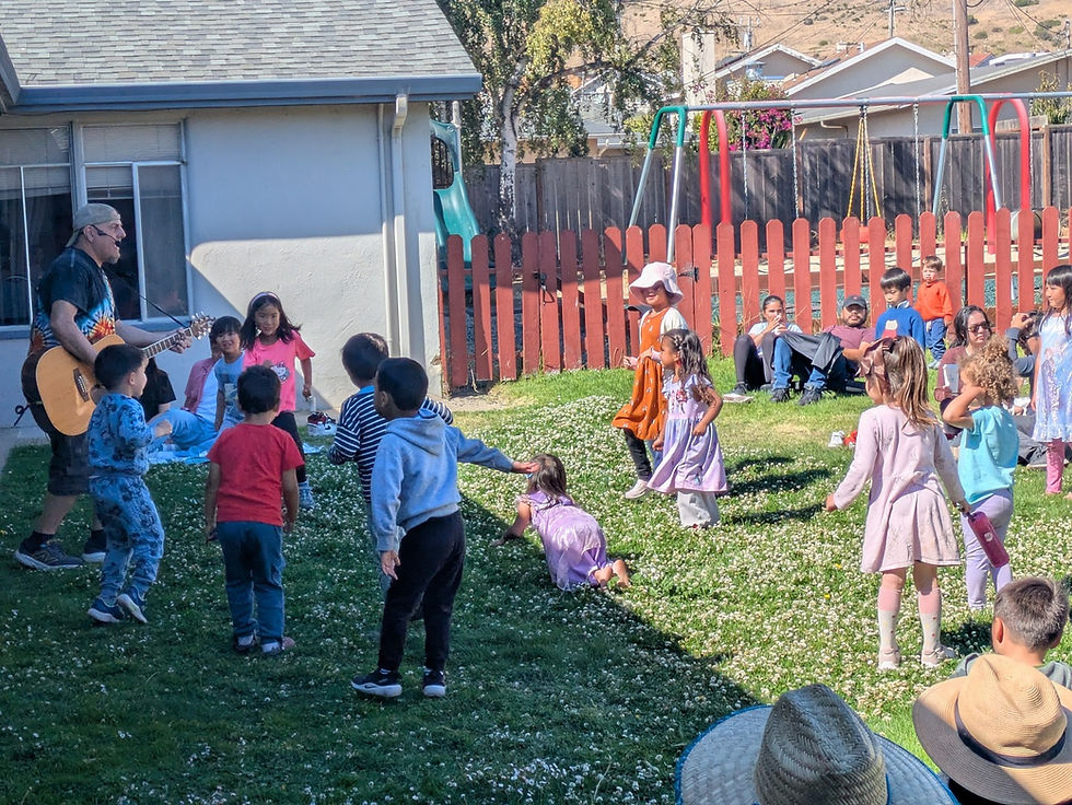 Children gather on a grassy playground while an adult plays guitar and families watch nearby.