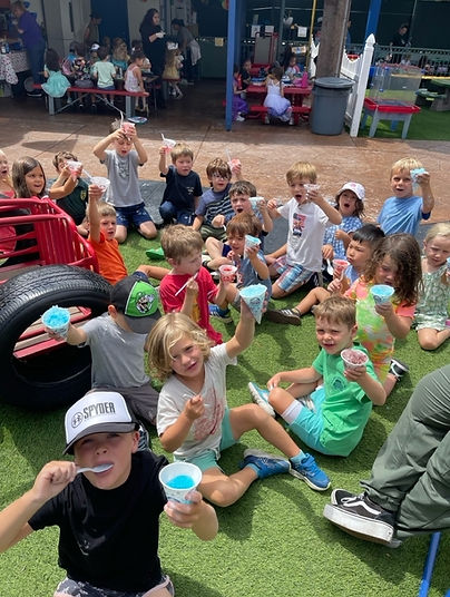 A group of young children sitting on artificial grass outdoors, smiling and holding up colorful cups of shaved ice or frozen treats, with a playground and picnic tables visible in the background.
