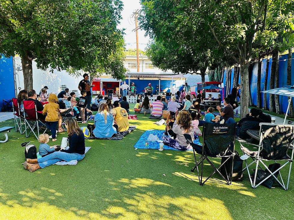 Families and children gather outdoors on blankets and lawn chairs under shady trees, watching a live performance in a grassy community play area.
