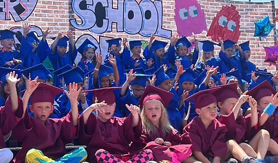 Large group of preschool graduates seated on outdoor risers during a graduation ceremony. Children in maroon and blue caps and gowns raise their hands while singing or participating in a performance. A colorful backdrop behind them features a brick-wall design, the word “SCHOOL,” and video game-inspired characters and decorations. Bright sunshine and festive balloons add to the celebratory atmosphere.