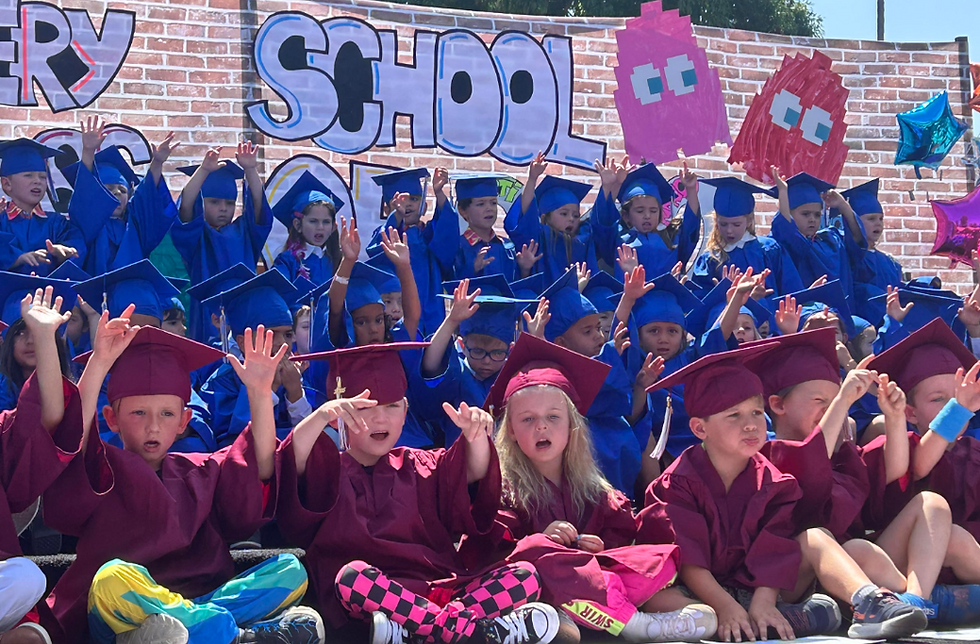 Children in blue and burgundy graduation caps and gowns perform on an outdoor stage during a preschool graduation ceremony, with a colorful “School” backdrop and decorations behind them.
