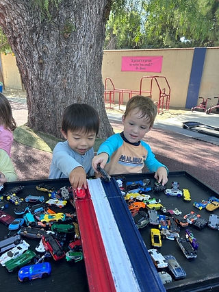 Two young children playing with toy cars on a colorful race track at an outdoor play area.