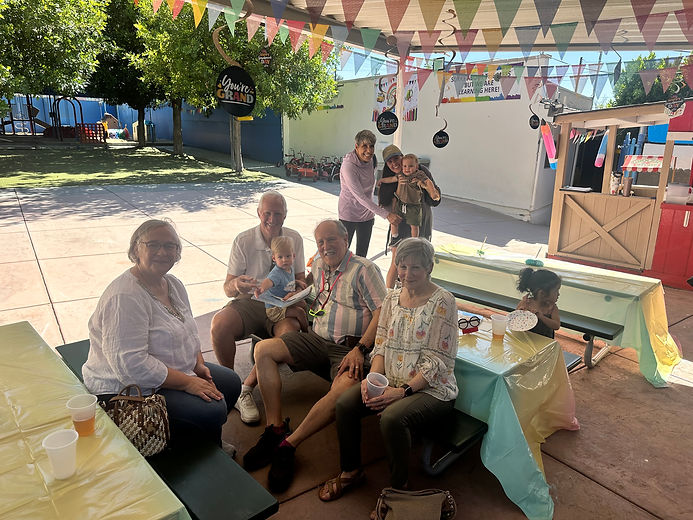 Grandparents gather at picnic tables under colorful bunting in a preschool courtyard, smiling together while two in the background hold young children.