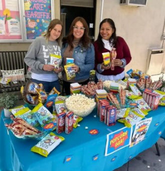 Three women stand behind a colorful popcorn bar table outdoors, smiling and holding snacks. The table is filled with popcorn, candy, and treats, and a sign behind them reads “Moms Are Tea-riffic,” creating a fun, celebratory event atmosphere.