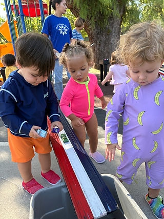 Three young children playing with toy cars on a colorful race track at an outdoor playground.