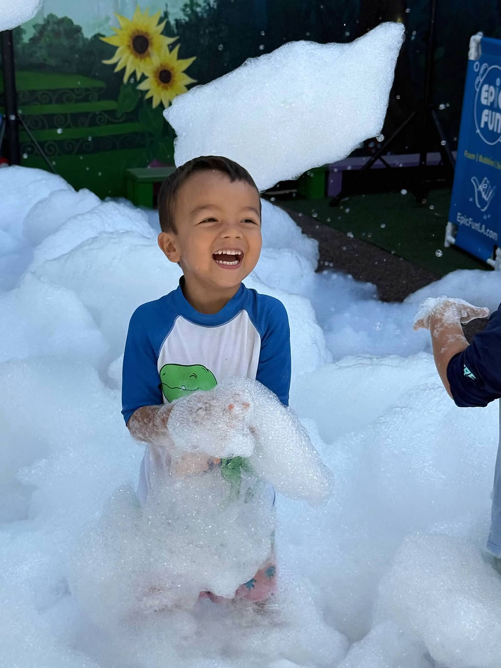 A laughing young child stands surrounded by fluffy white foam during a bubble play activity. Wearing a blue-and-white swim shirt, the child holds a mound of bubbles while foam floats through the air and piles up around them. The outdoor play area features colorful decorations in the background, creating a joyful and playful summer scene.