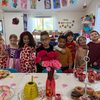 Seven young children stand arm in arm and smile in a classroom decorated for Valentine’s Day, with a table of heart-themed treats in the foreground, including pink flowers, cookies, donuts, and cups of strawberries.